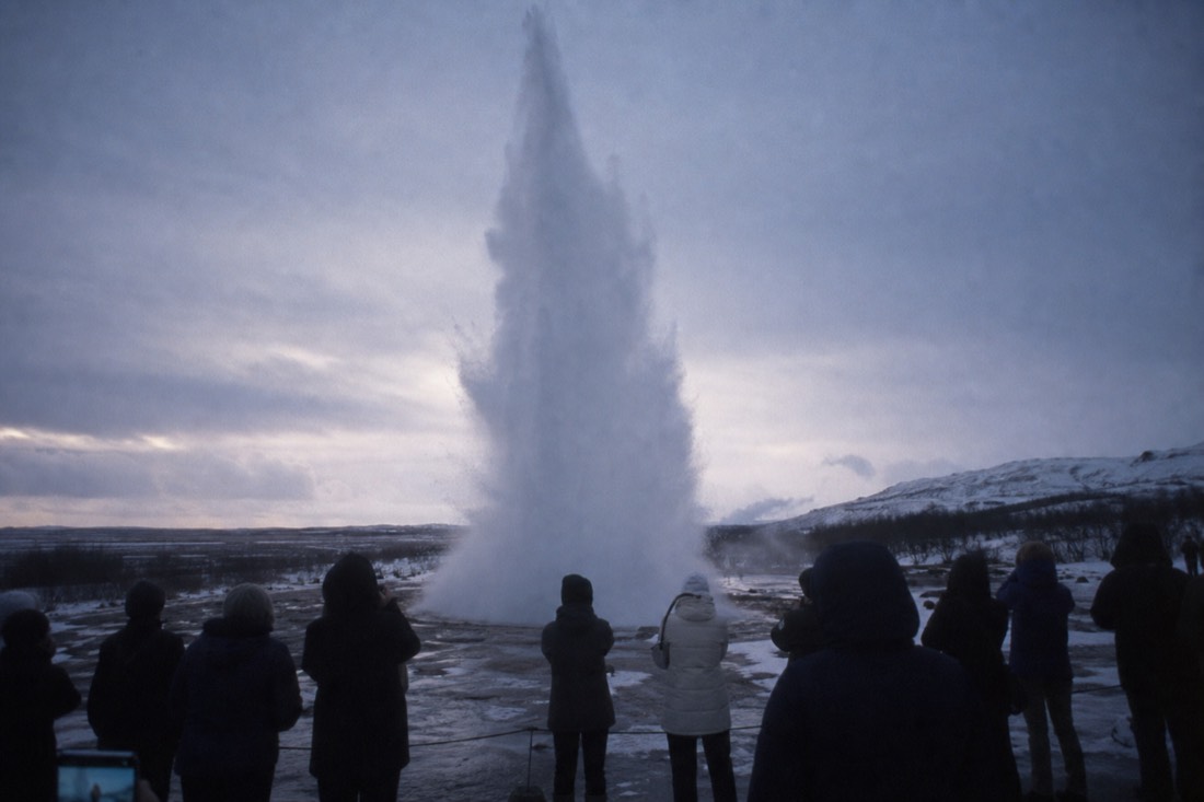 Strokkur geyser erupting in a tall white column at winter dusk with crowd silhouettes at the rope line