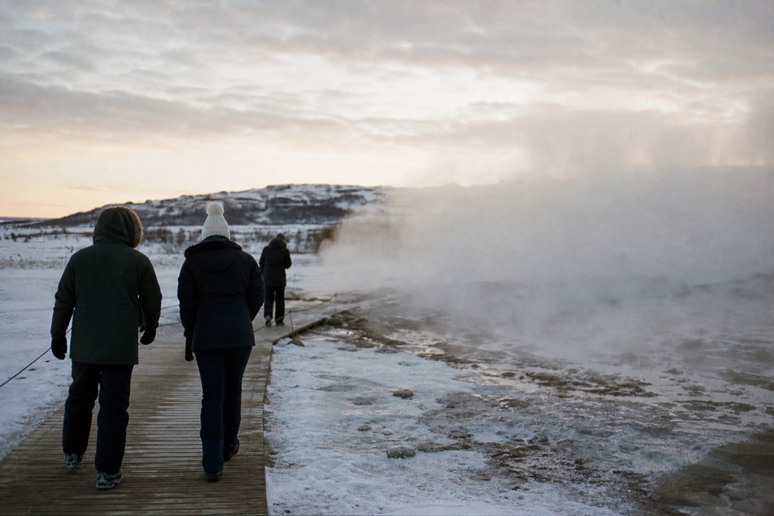 Visitors in winter parkas seen from behind on the Geysir boardwalk, steam blowing across packed snow