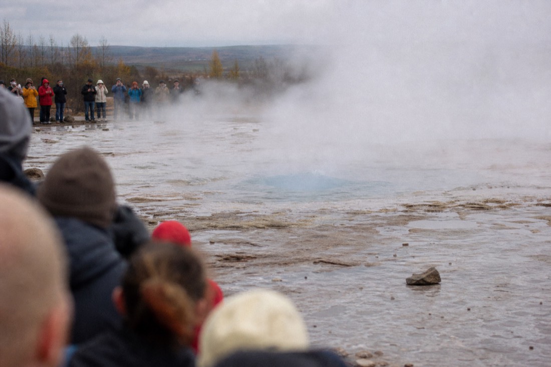 Crowd waiting around the roped edge of Strokkur at Geysir as steam drifts across the field