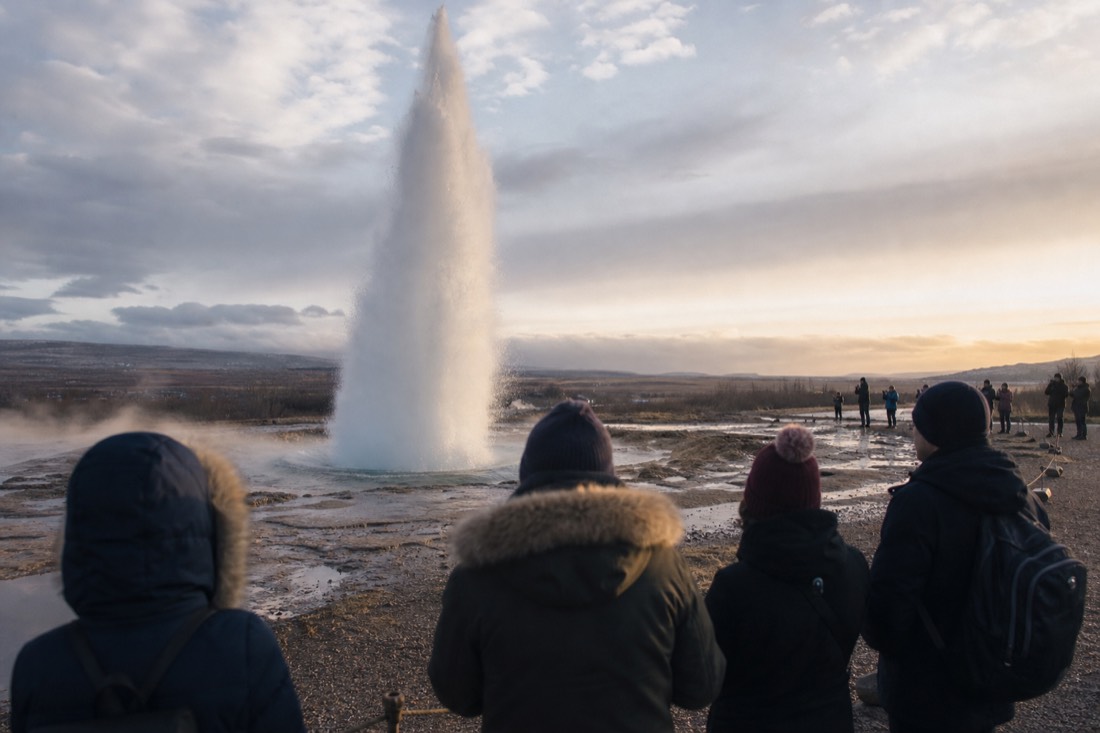 Strokkur geyser mid-eruption with onlookers seen from behind on the roped path