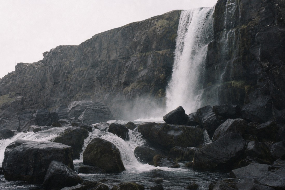 Öxarárfoss waterfall dropping over the dark basalt wall of Almannagjá at Þingvellir with spray over the boulder field below